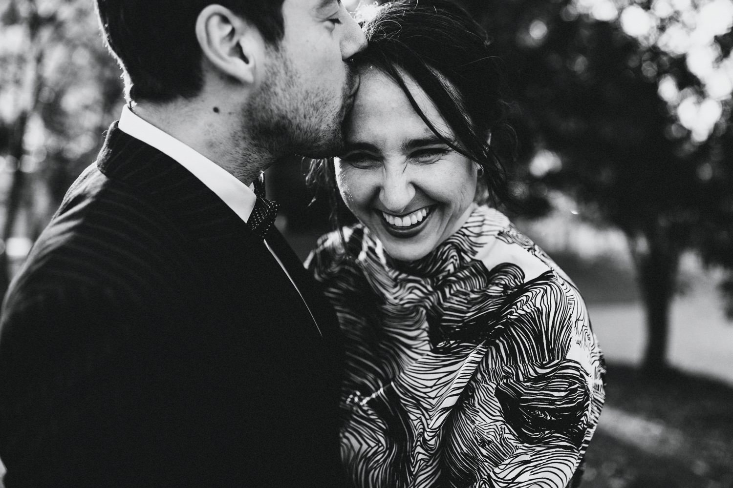 Der Bräutigam gibt seiner Braut einen zärtlichen Kuss auf die Schläfe, während die Braut herzhaft lacht und sich liebevoll an ihn kuschelt. In diesem intimen Moment der Zuneigung und Verbundenheit strahlen sie vor Glück und Vorfreude auf ihre gemeinsame Zukunft.