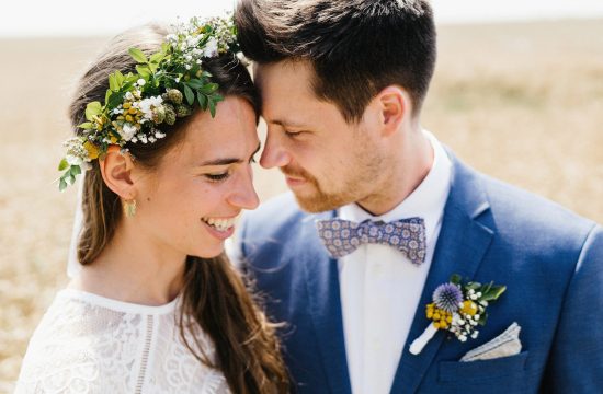 Ein Brautpaar kuschelt innig in einem Weizenfeld die Braut trägt ein Blumenkranz der Bräutigam einen blauen Anzug und eine Schleife
