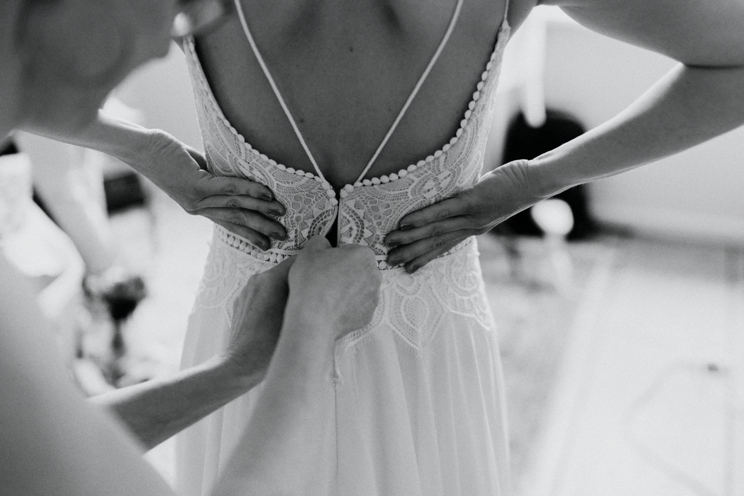 Die Hände der Freundin der Braut helfen der Braut beim Schließen des Reißverschlusses des Brautkleides mit schöner Spitze. In diesem berührenden Moment sieht man nur die liebevollen Gesten der Unterstützung, während sich die Braut auf ihren großen Tag vorbereitet. Der Rücken der Braut strahlt Eleganz und Vorfreude aus, während ihre Freundin behutsam dafür sorgt, dass das Kleid perfekt sitzt.