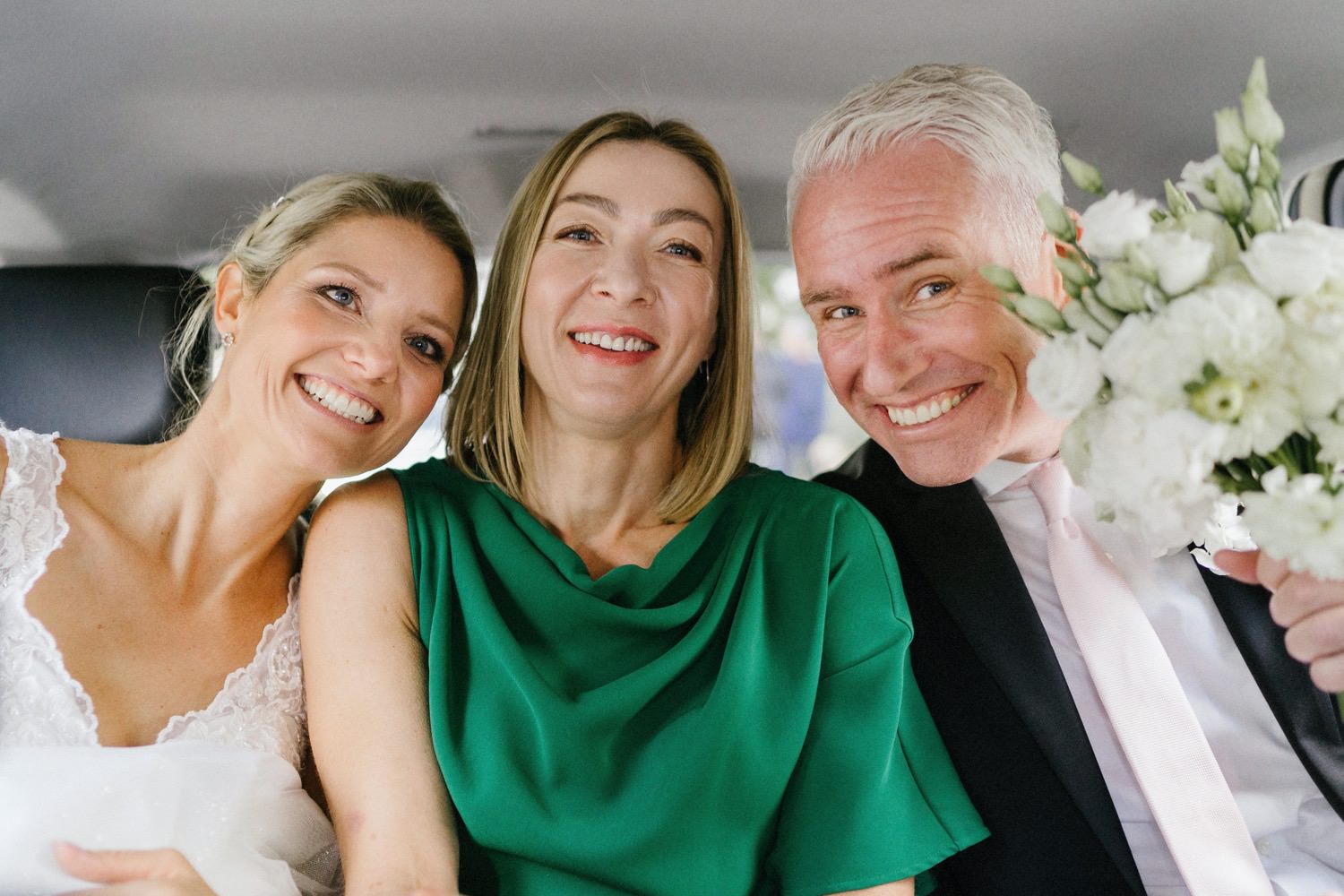 Das Brautpaar fährt mit einer Trauzeugin in der Mitte auf der Rückbank eines Taxis zur Feierlocation im The Fontenay