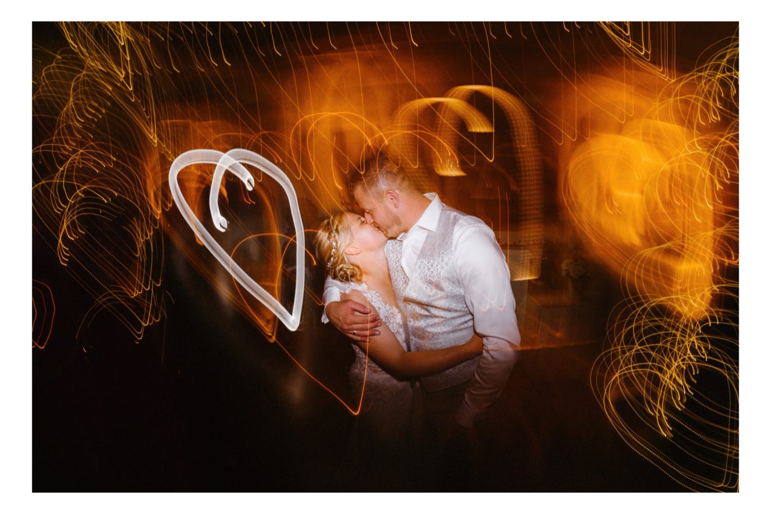 Das Brautpaar versinkt in einem verträumten Kuss auf der Tanzfläche, während der Hochzeitsfotograf mit einer Langzeitbelichtung ein strahlendes Herz um sie herum malt. Das Bild erfasst die Intimität und Romantik des Moments, während das leuchtende Herz die tiefe Liebe und Verbundenheit des Paares symbolisiert. Die sanfte Bewegung der Tanzenden und das warme Licht der Location verstärken die zauberhafte Atmosphäre und machen diesen Augenblick zu einem unvergesslichen Höhepunkt der Feier. Es ist ein Bild voller Leidenschaft und Zuneigung, das die Magie der Liebe perfekt einfängt und für die Ewigkeit festhält.