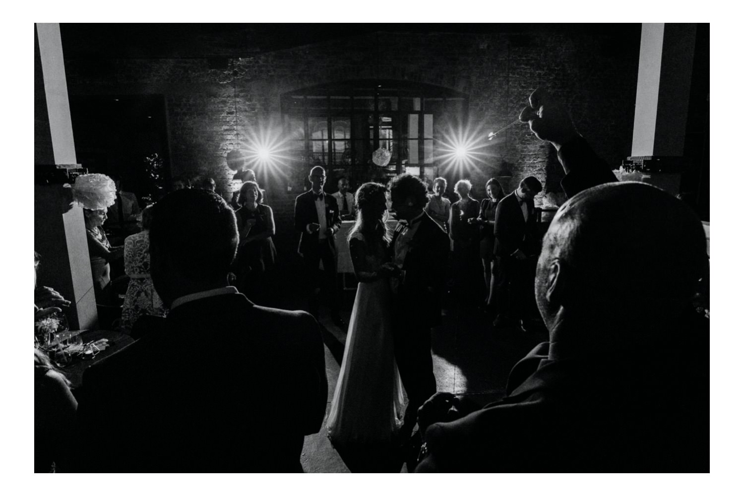 Das Brautpaar tanzt im Mittelpunkt des Geschehens, umgeben von den lachenden Gesichtern der Gäste. Das Gegenlicht der Blitze verleiht der Szene eine magische Atmosphäre, während das Brautpaar den Moment des ersten Tanzes in vollen Zügen genießt.